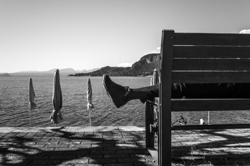 Dell'uomo sdraiato sulla panchina si vedono i piedi, sullo sfondo il lago di Garda. Street Photography, Black and White.