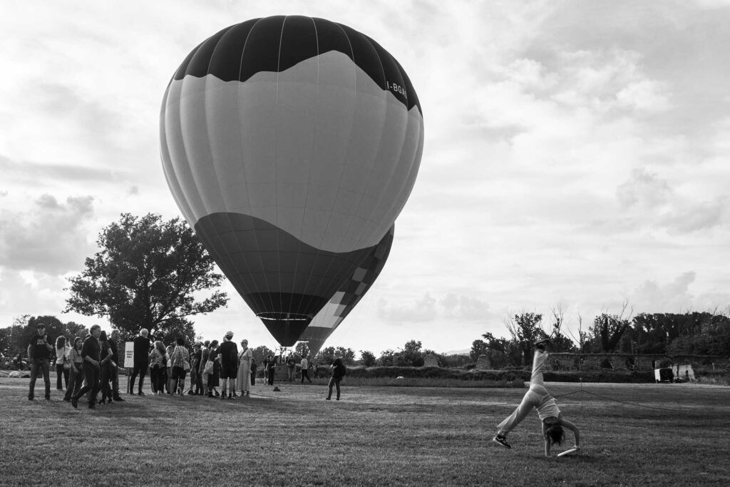 Mongolfiere in partenza sul prato mentre una bambina fa le capriole. Street Photography, Black and White.