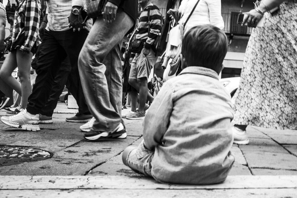 Un bambino seduto sulla via osserva dalla sua altezza le gambe dei passati. Street Photography, Black and White.