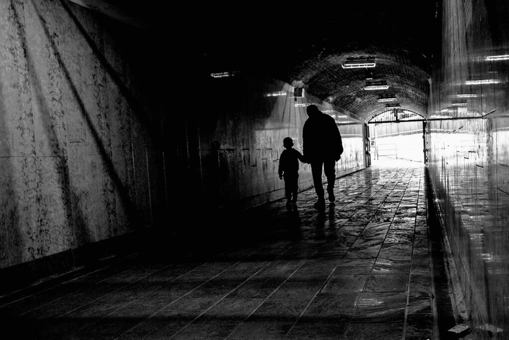 Un bambino stringe la mano al papà mentre attraversa il buio di una galleria. Street Photography, Black and White.