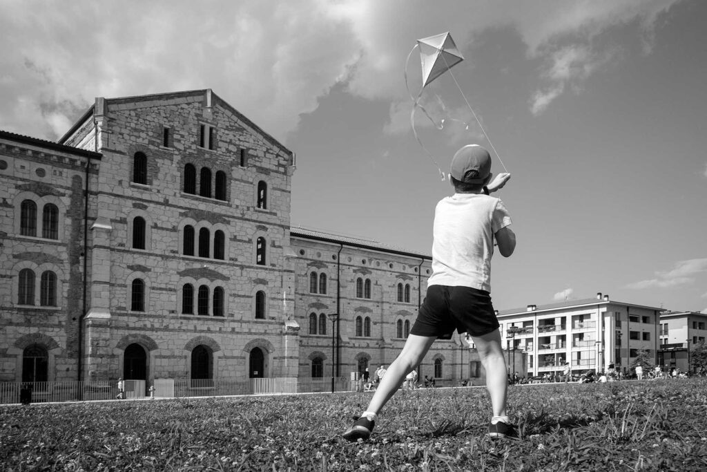 Un ragazzo e il suo aquilone in un prato tra palazzi antichi e moderni. Street Photography, Black and White.