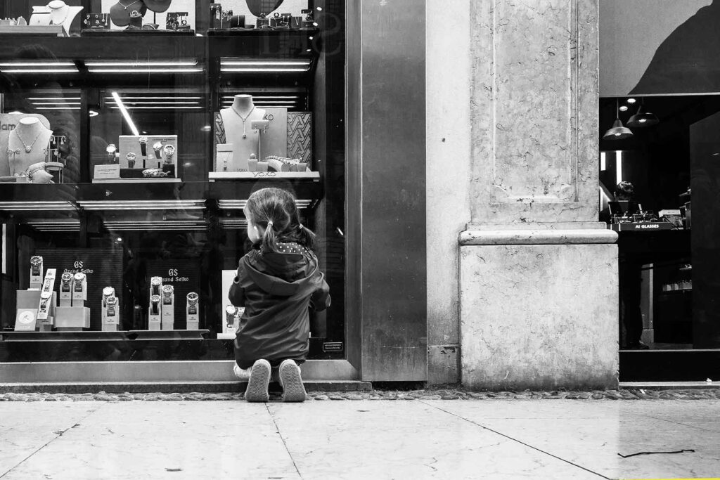 Bambina in ginocchio rapita dagli oggetti presenti in vetrina. Street Photography, Black and White.