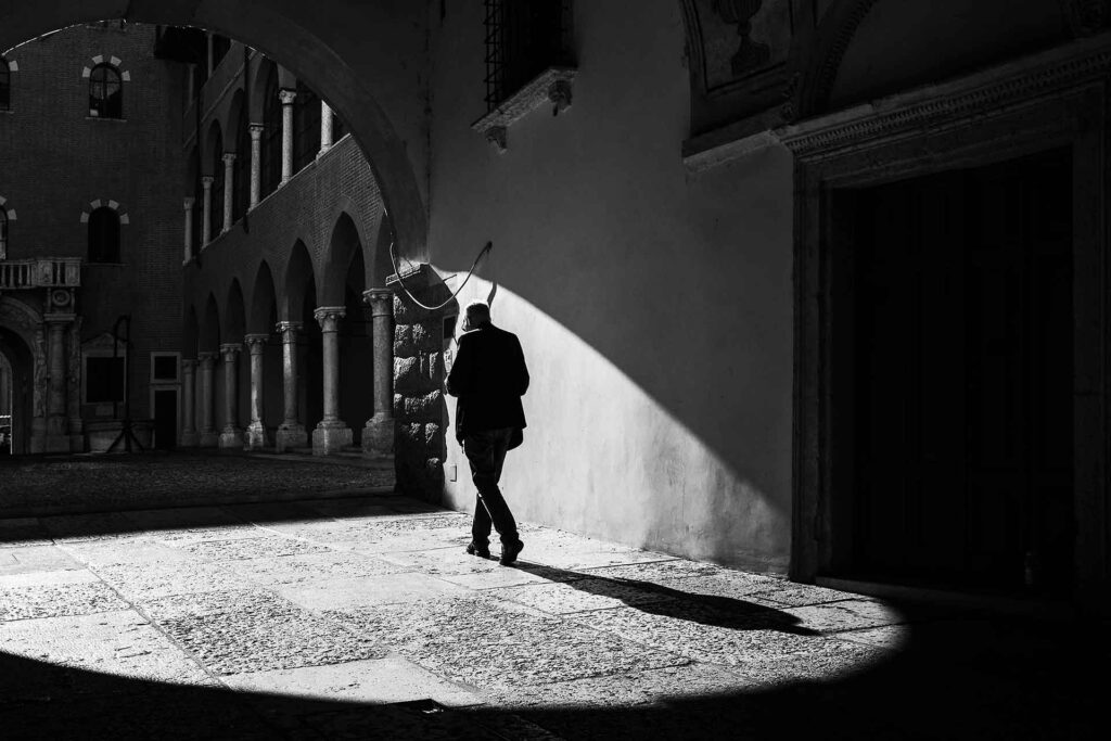 Un uomo sotto il volto di una piazza antica proietta l'ombra sul pavimento in pietra. Street Photography, Black and White.
