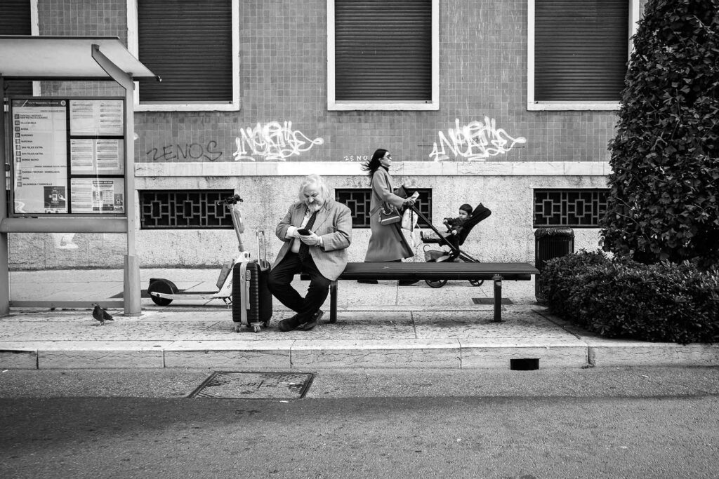 Un signore ben vestito in attesa del bus mentre passa una signora con il suo bambino. Street Photography, Black and White.