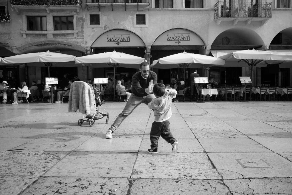 Padre e figlio giocano sulla piazza e si prendono la scena: "Dammi un cinque!". Street Photography, Black and White.