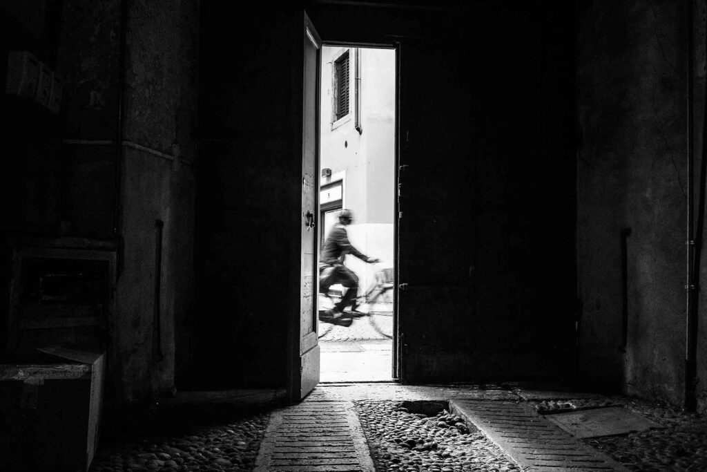 Da un androne il portone semiaperto mostra un ciclista di passaggio. Street Photography, Black and White.