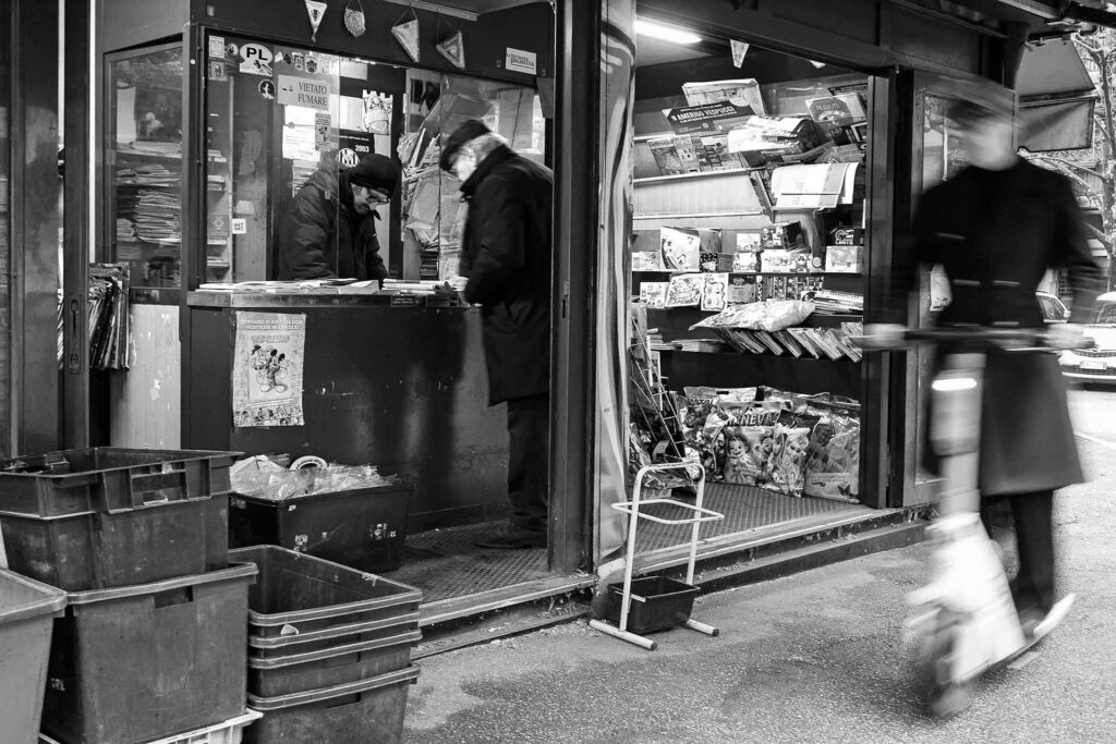 Un signore compera il giornale all'edicola mentre sfreccia un monopattino. Street Photography, Black and White.