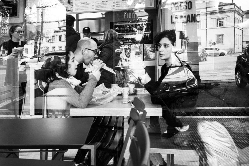 Una famiglia in un bar consuma il pasto tra i riflessi della vetrina. Street Photography, Black and White.