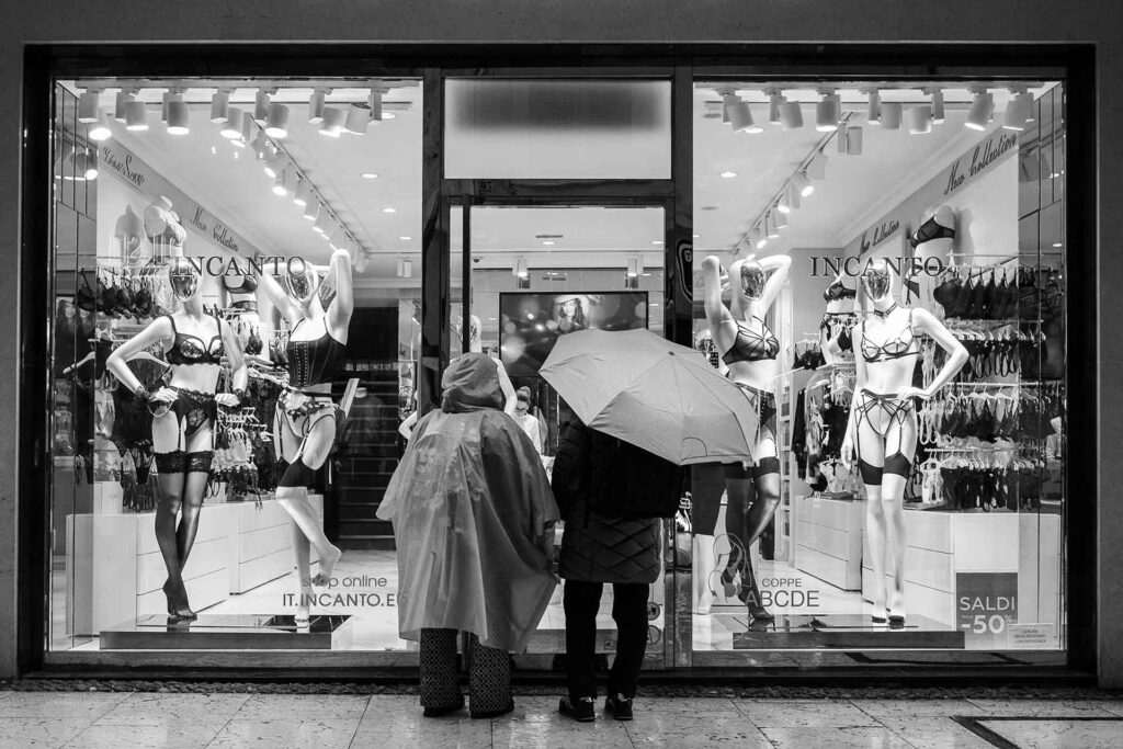 Piove, una coppia anziana osserva la vetrina che espone lingerie. Street Photography, Black and White.