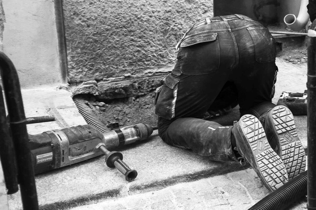 Il sedere di un operaio che ha la testa immersa nel buco dove sta lavorando. Street Photography, Black and White.