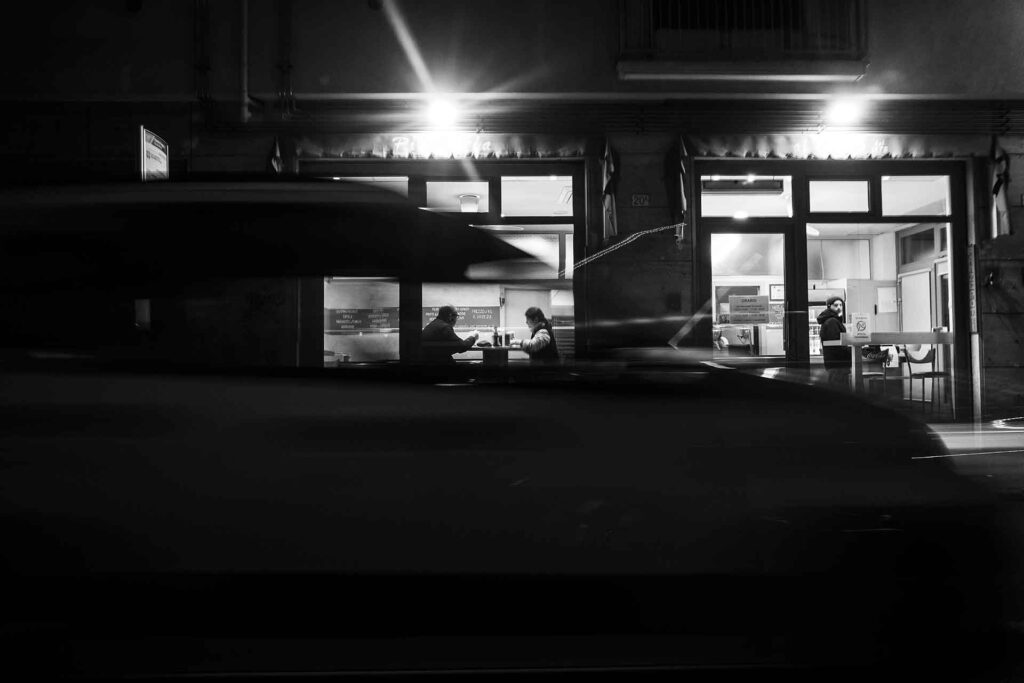 Foto di strada notturna: due persone cenano dietro una vetrina illuminata. Street Photography, Black and White.