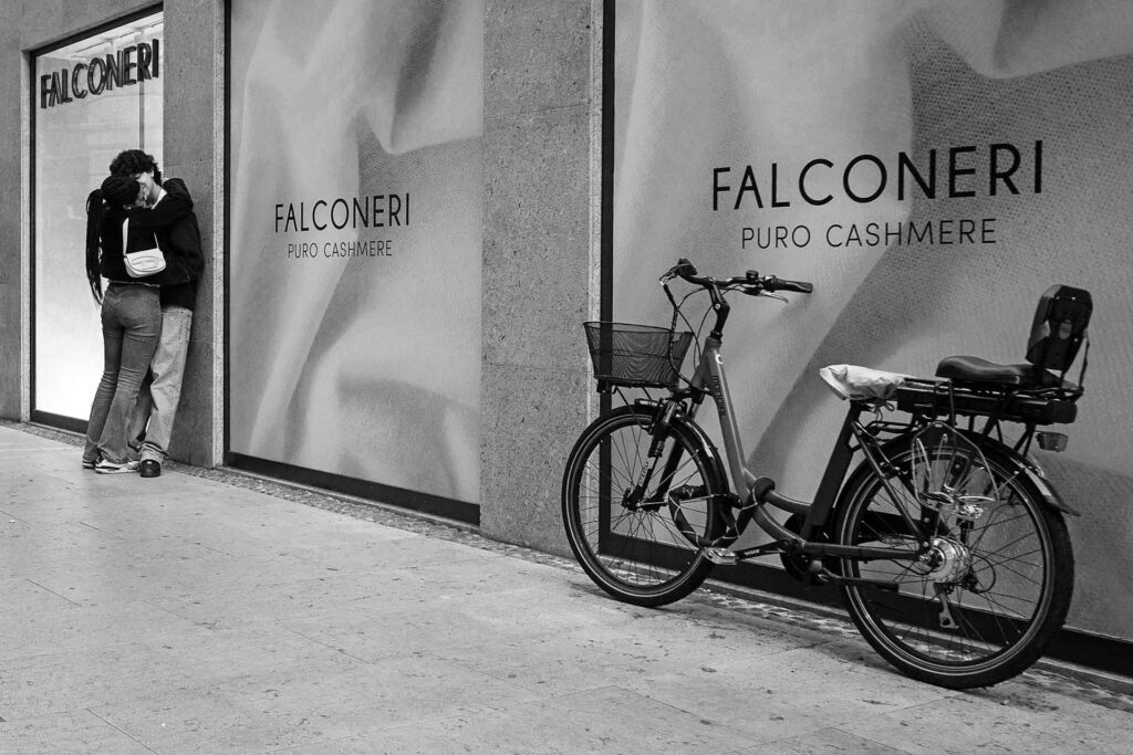 Un ragazzo e una ragazza abbracciati, appoggiati a un muro con una bicicletta in primo piano. Street Photography, Black and White.