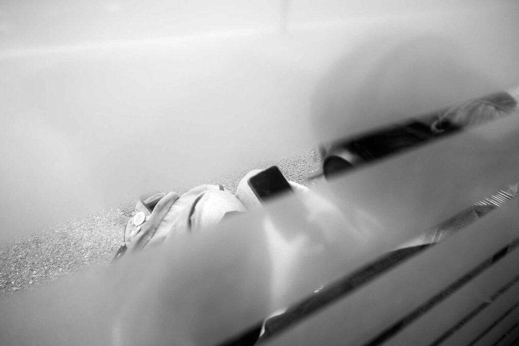 Una ragazza con le cuffie e il telefonino seduta sul marciapiede appoggiata a una vetrina. Street Photography, Black and White.