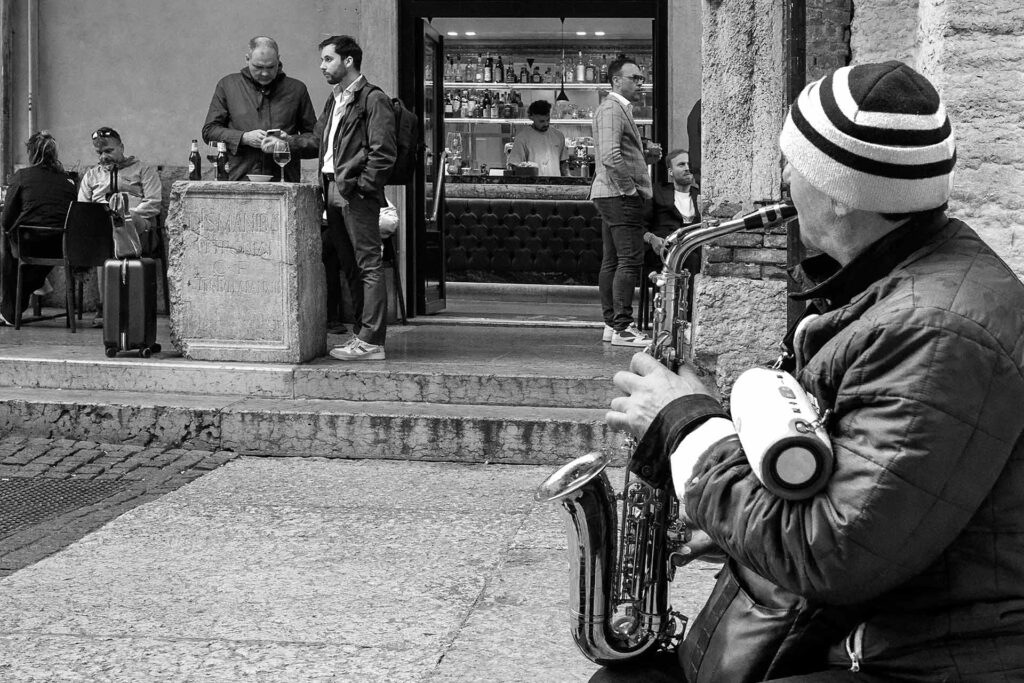 Un artista di strada suona il sax mentre osserva la vita scorrere dentro il bar. Street Photography, Black and White.